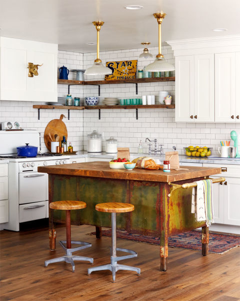 A wood and metal military desk-turned-prep island is the centerpiece of this Arizona ranch home's kitchen. The weathered wood top and rusty green base give the kitchen more personality. Barnwood shelves pick up the warmth of the island's counter, while brass pendants (from an old ice-cream parlor) echo the towel bar's shine. 